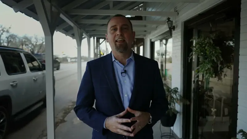 Adam Simmons in a navy blazer, speaking outside a storefront with a covered porch